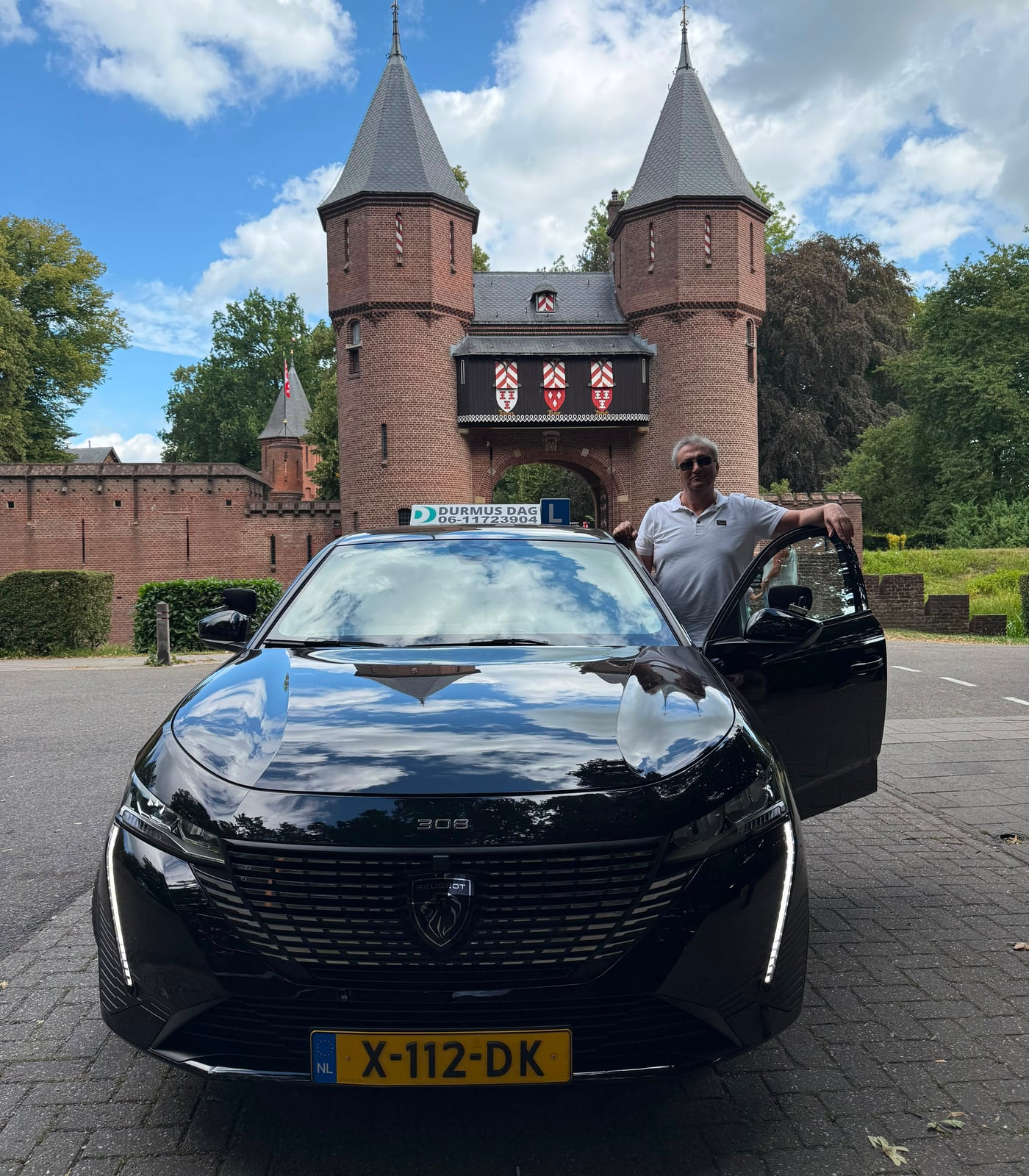 Rij-instructeur Durmus Dag naast de geopende deur van de zwarte Peugeot-lesauto, voor het poortgebouw van Kasteel de Haar